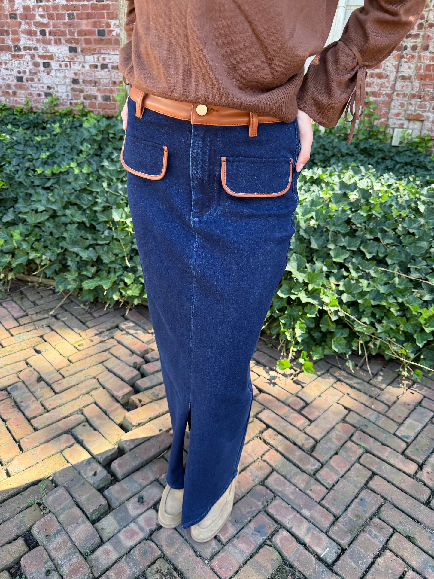 Close view of blue denim maxi skirt with chestnut leather trim detail, front flap pockets and zipper closure slit