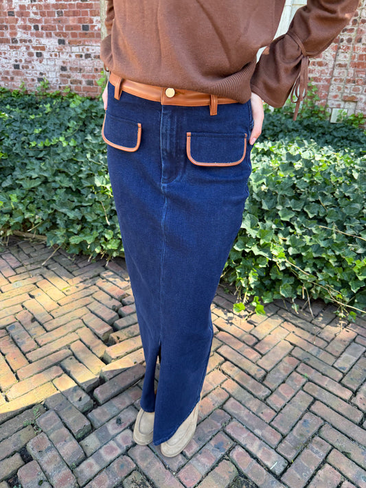 Close view of blue denim maxi skirt with chestnut leather trim detail, front flap pockets and zipper closure slit