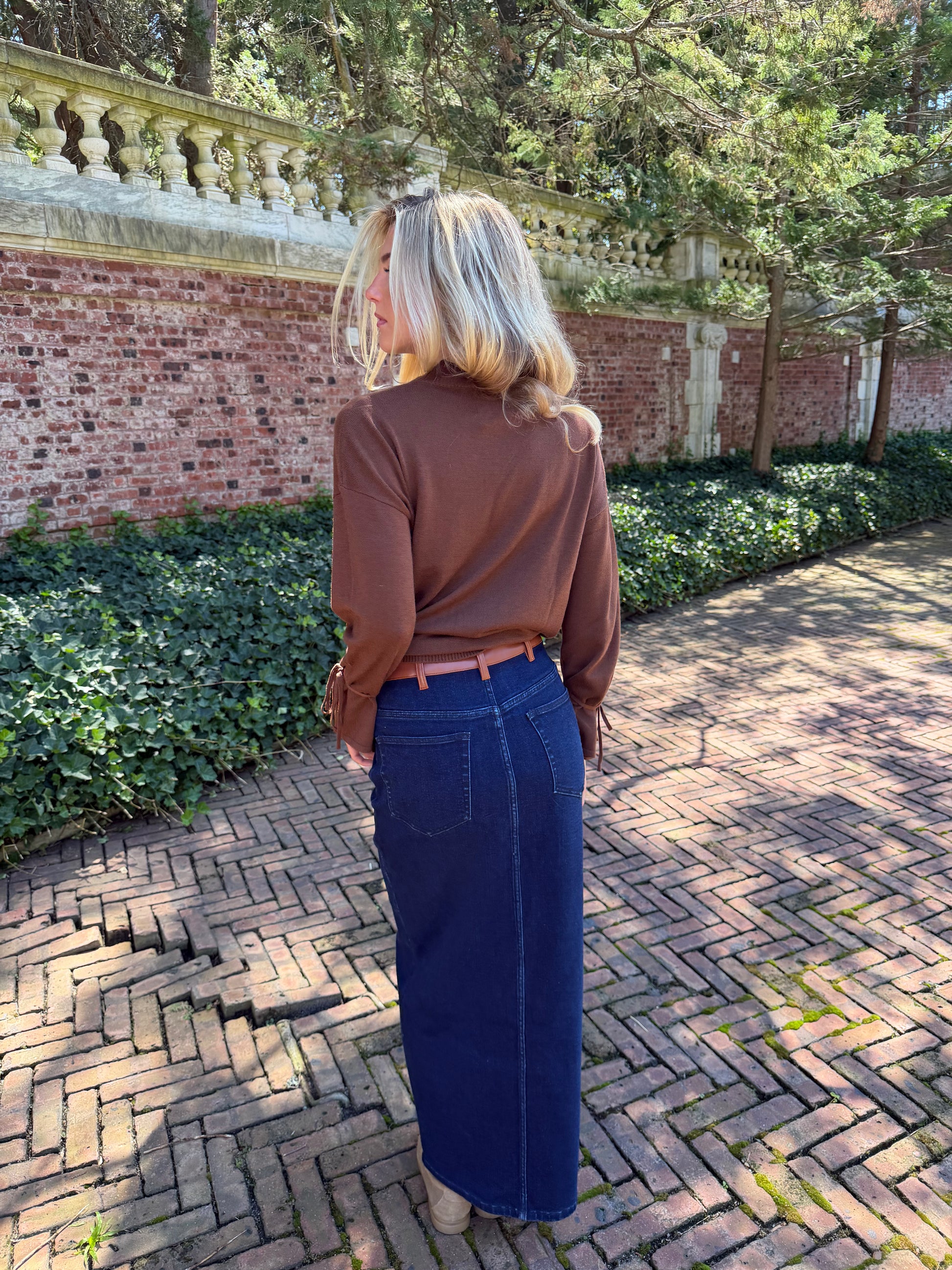 Back view of blue denim maxi skirt with chestnut leather trim detail, front flap pockets and zipper closure slit