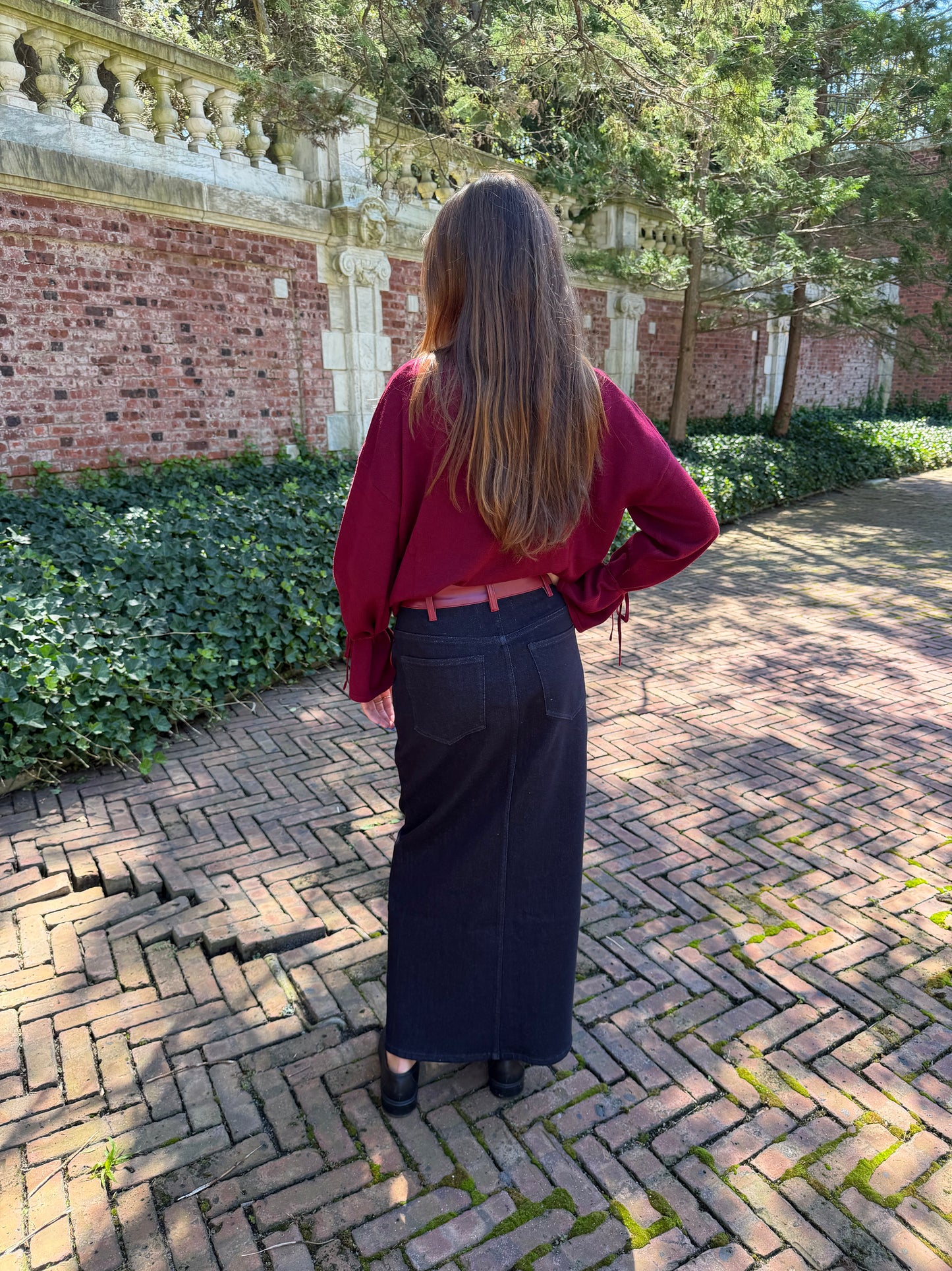 Back view of black denim maxi skirt with burgundy leather trim detail, front flap pockets and zipper closure slit