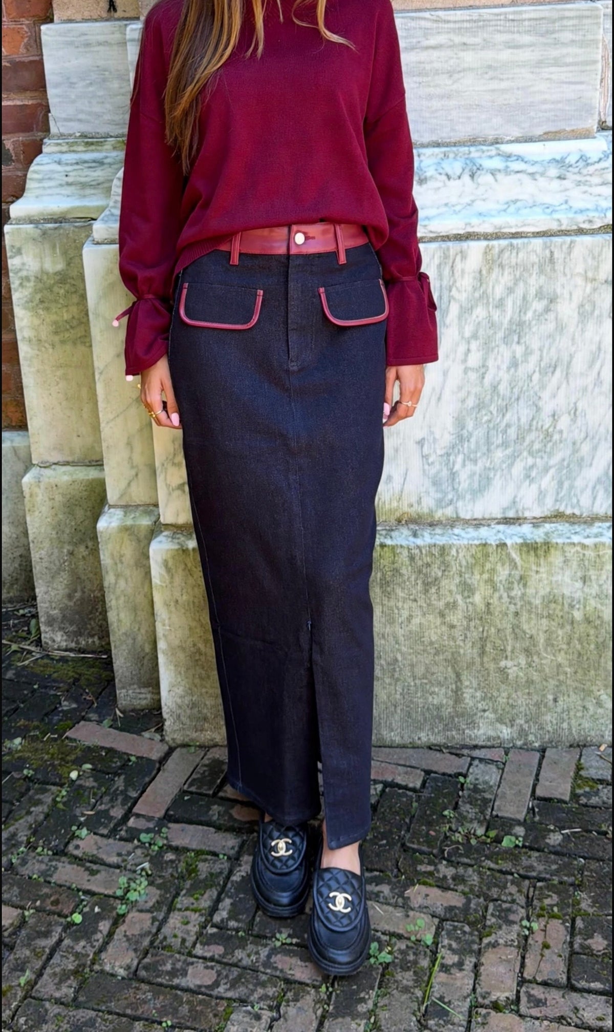 Front view of black denim maxi skirt with burgundy leather trim detail, front flap pockets and zipper closure slit